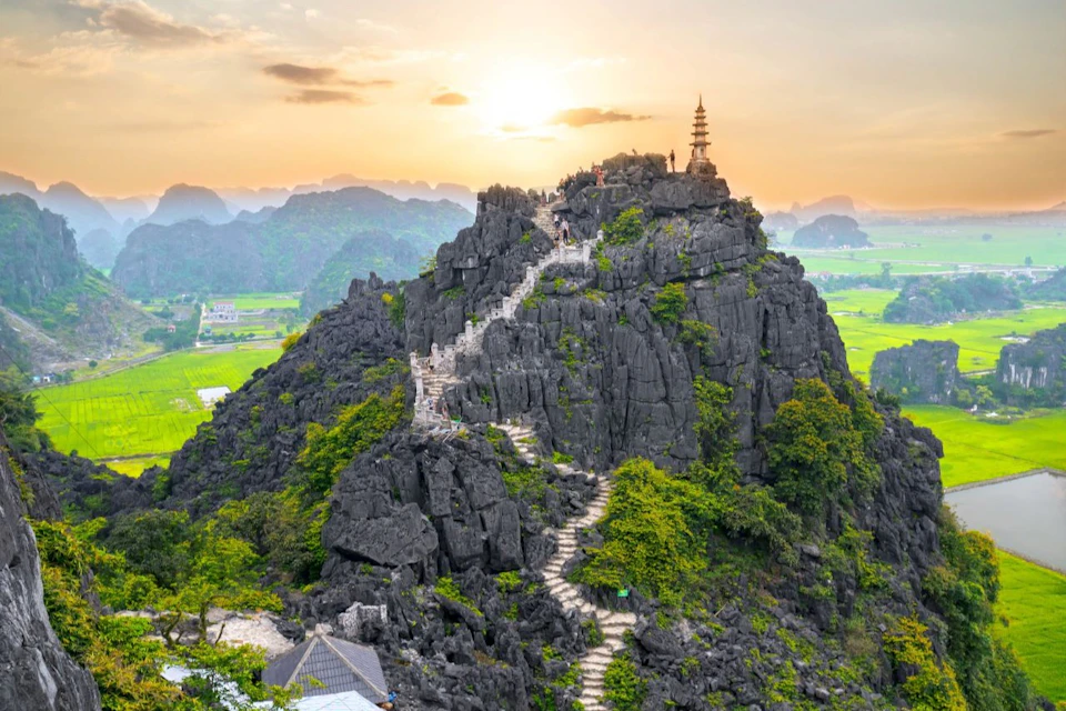 Stone staircase winding up a rocky, green hill towards a small pagoda at the summit with a scenic landscape in the background.