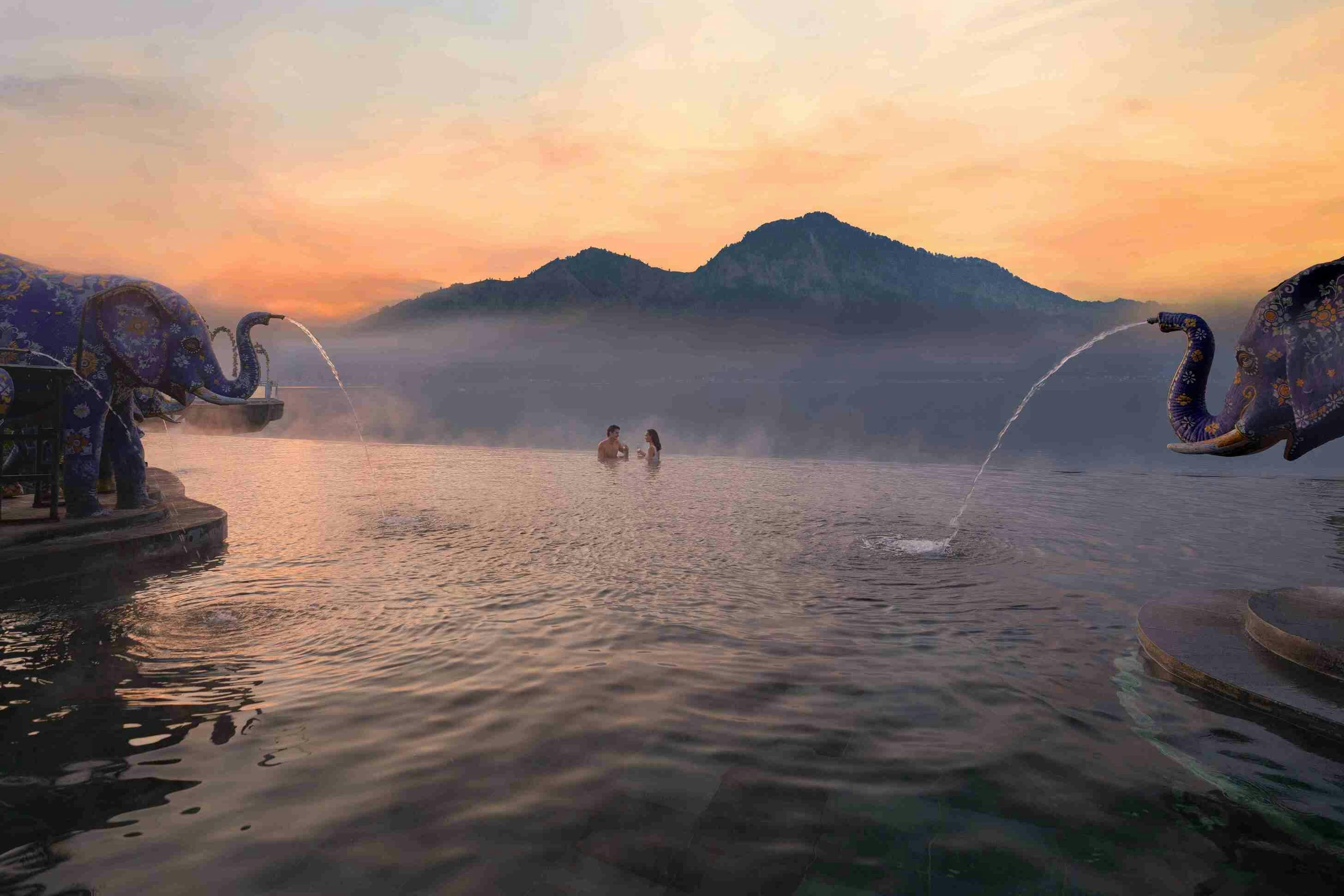 Two people standing in a calm infinity pool with mountain scenery and water fountains under a colorful sky at sunset.