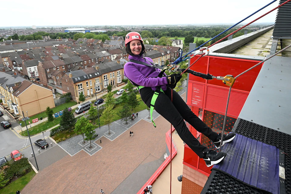 Woman in helmet and harness smiles while abseiling down a red building; residential neighborhood and greenery in the background.