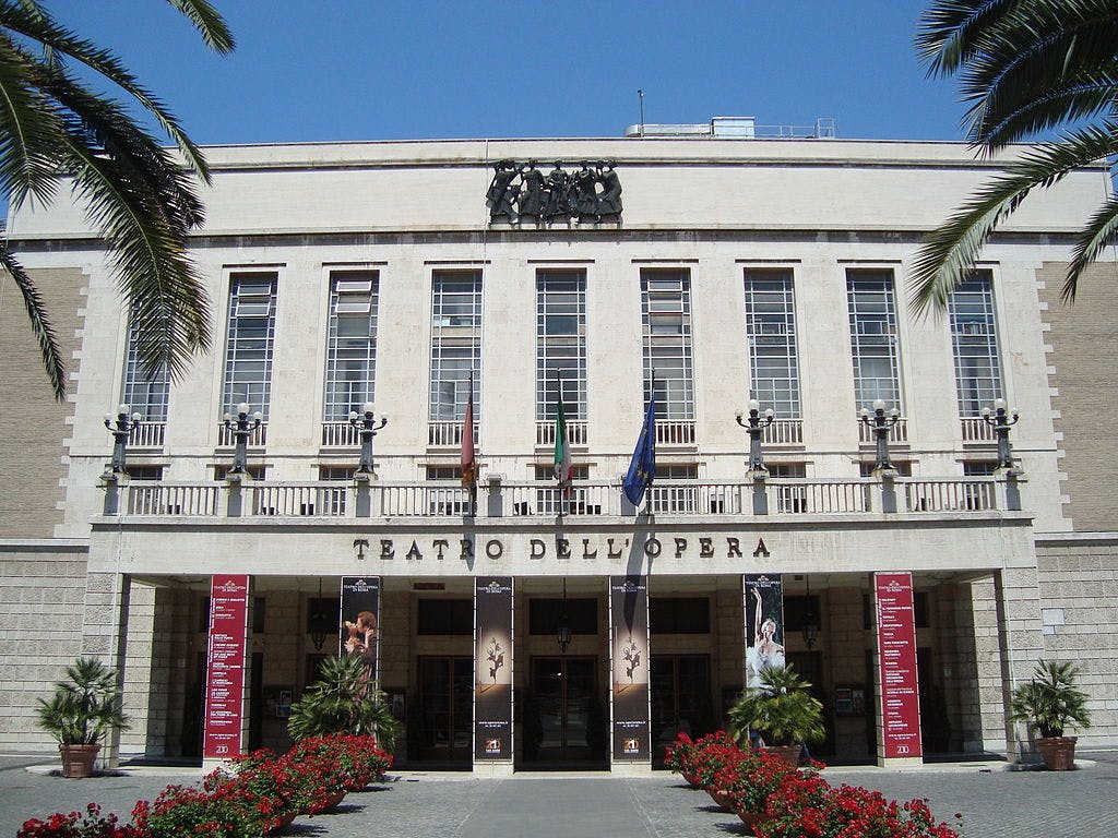 Façade du Teatro dell'Opera, Rome ; hautes fenêtres, grande entrée avec colonnes, drapeaux et bannières rouges. Des palmiers et des fleurs rouges encadrent la vue.
