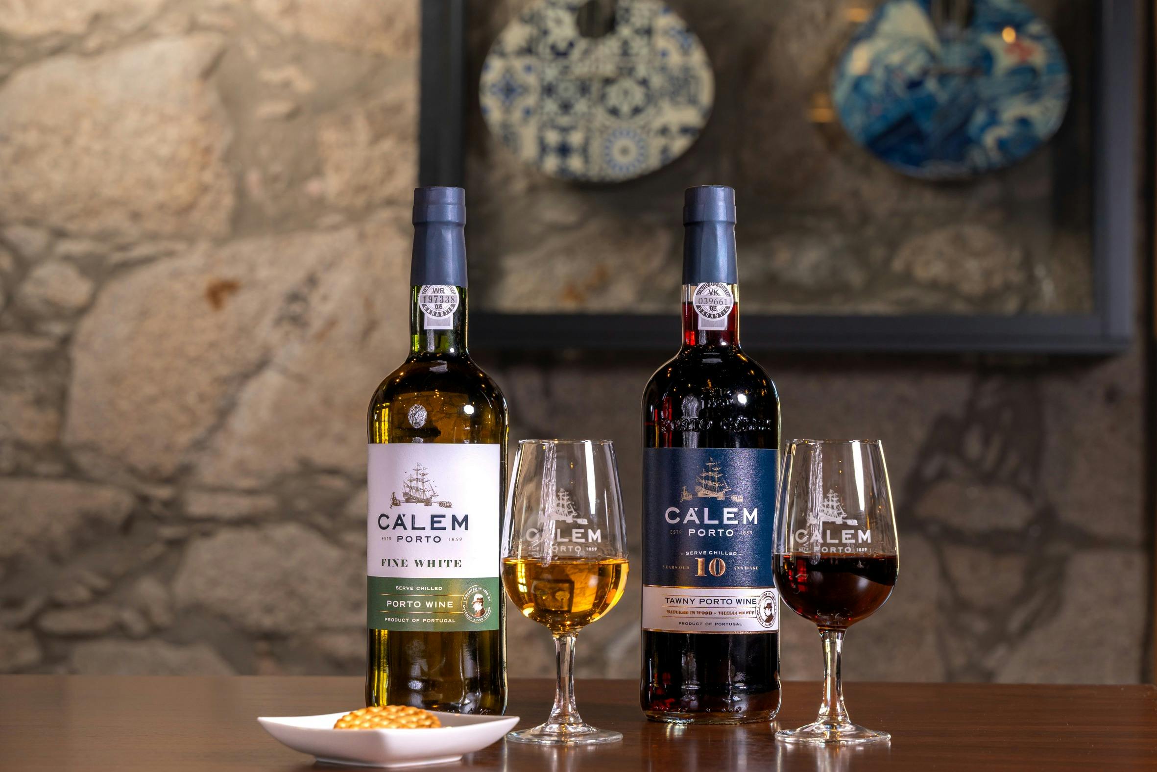 Two bottles of Calem Porto wine with glasses of white and red wine on a table, against a stone wall with decorative plates.