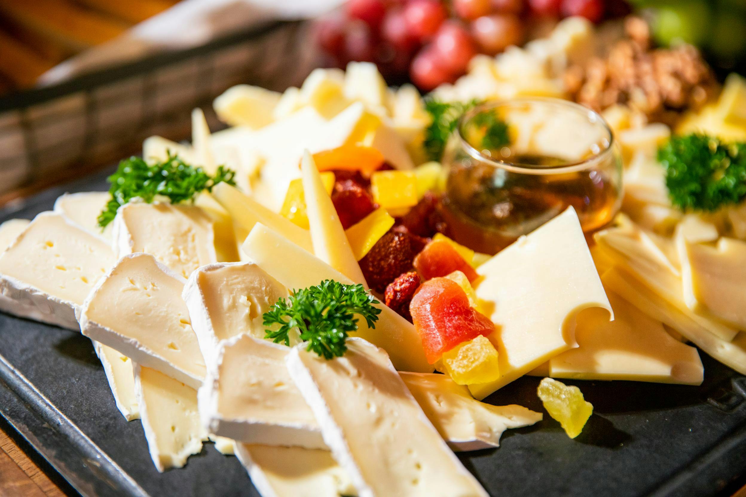 A close-up of a cheese platter featuring assorted cheeses, garnished with parsley, fruits, and a small container of honey.
