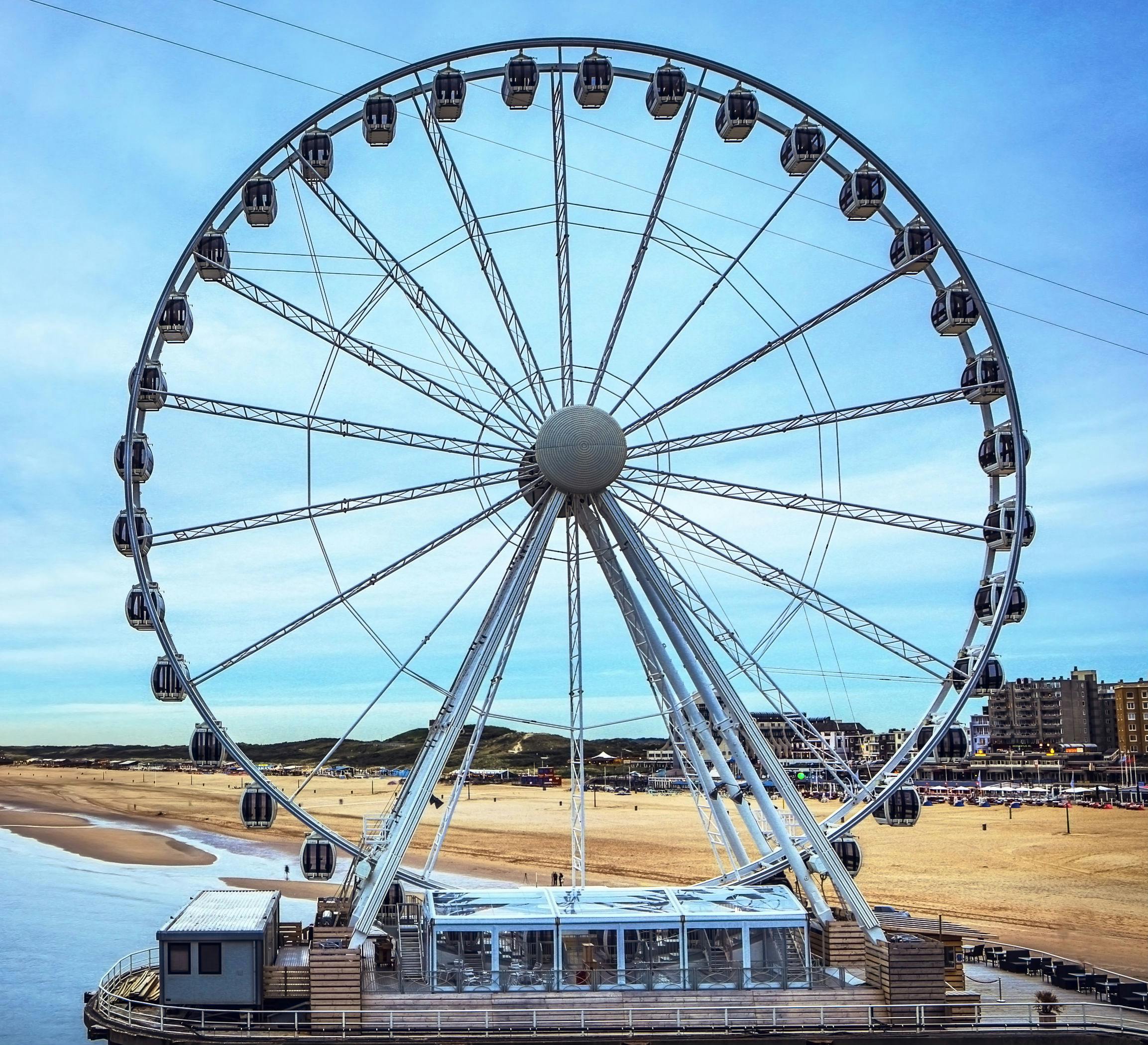 Velké ruské kolo na molu u písečné pláže s budovami a jasně modrou oblohou v pozadí.