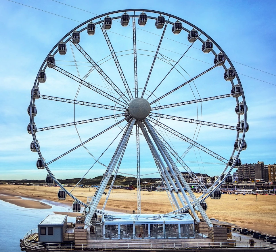 Large Ferris wheel on a pier by a sandy beach with buildings and a clear blue sky in the background.