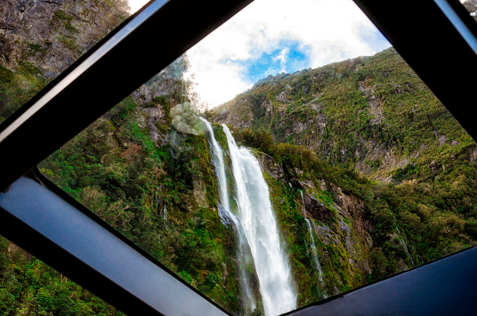 A waterfall flows down a green mountainside, viewed through a geometric window frame under a partly cloudy sky.