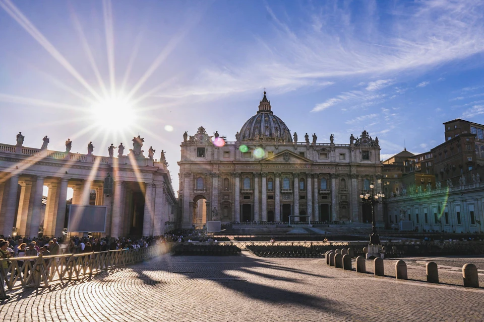 A view of St. Peter's Basilica in Vatican City under a clear blue sky with sunlight casting shadows on the plaza.