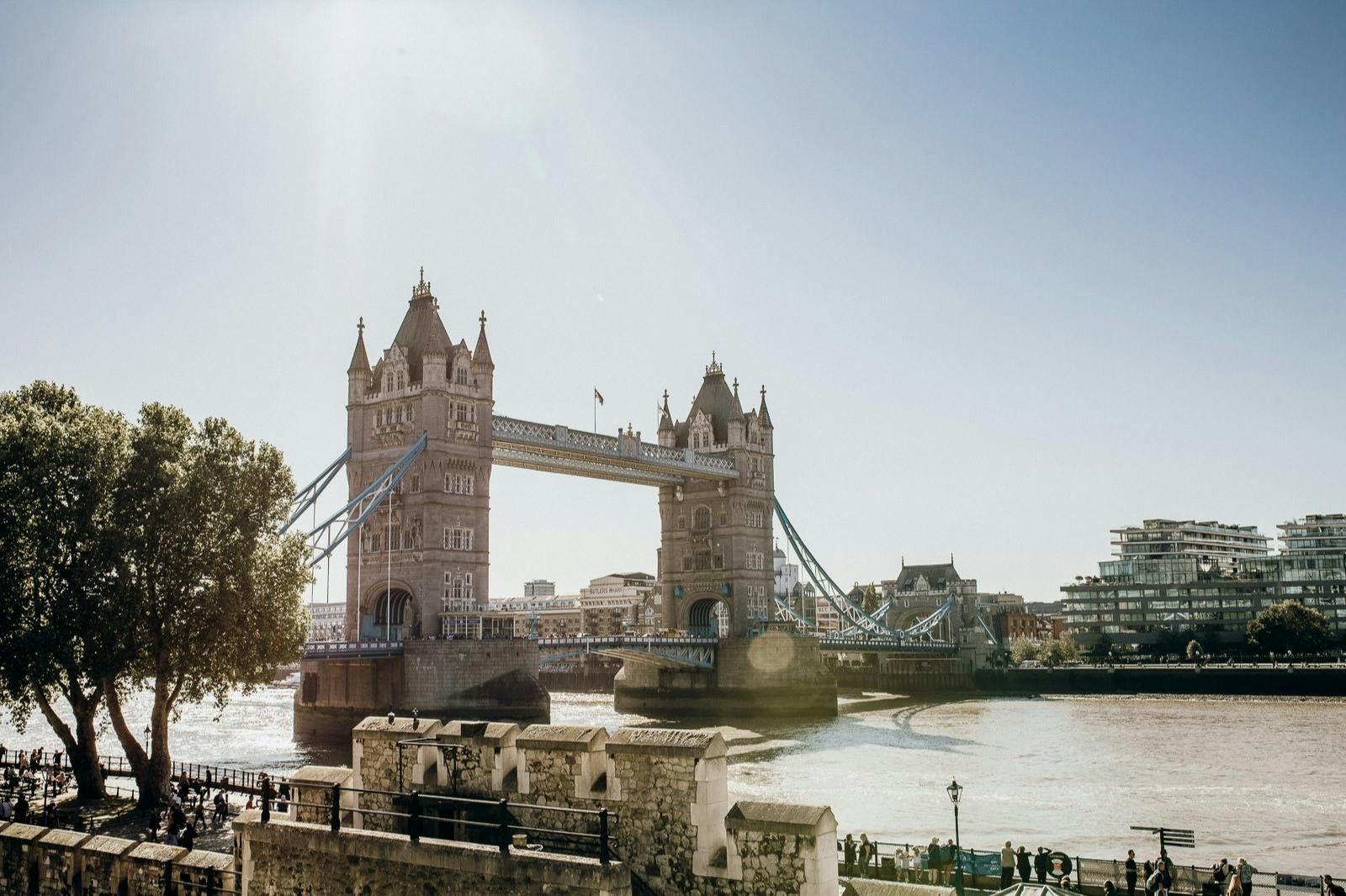 Tower Bridge of London spanning the River Thames on a sunny day, with people walking along the riverside.