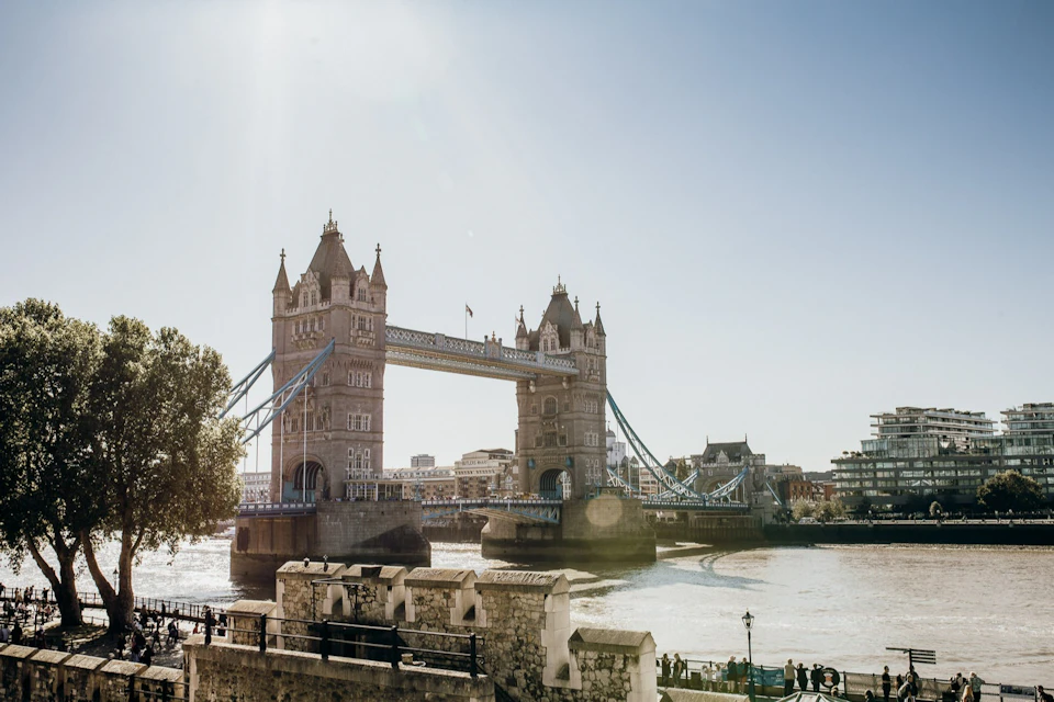Tower Bridge of London spanning the River Thames on a sunny day, with people walking along the riverside.