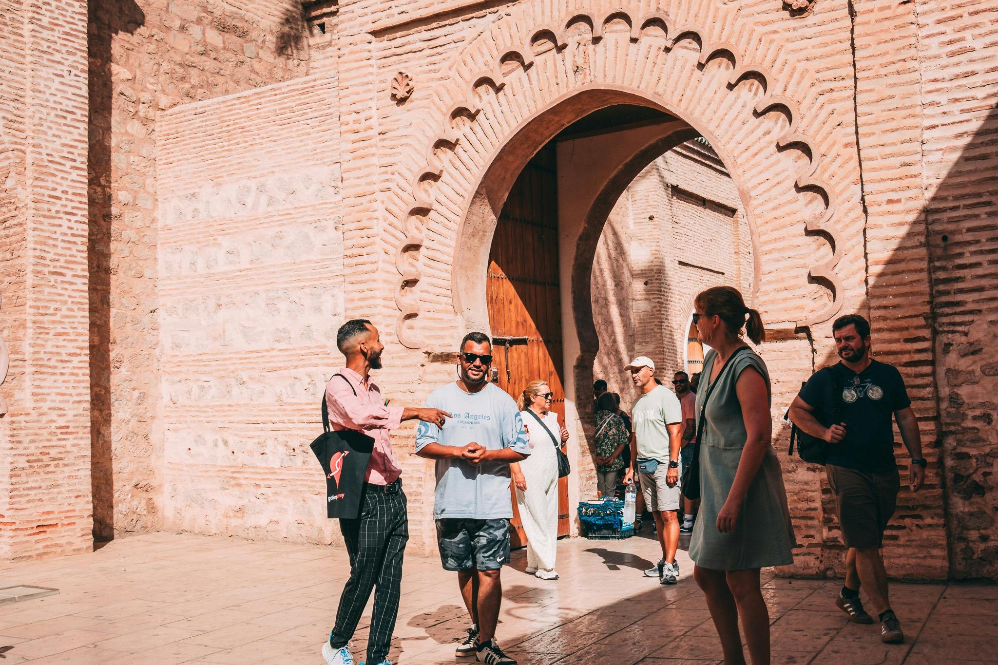 Οι άνθρωποι αλληλεπιδρούν και περπατούν κοντά σε μια μεγάλη, περίτεχνη πέτρινη καμάρα μια ηλιόλουστη μέρα. Ορισμένοι κρατούν τσάντες και φορούν καθημερινά ρούχα.