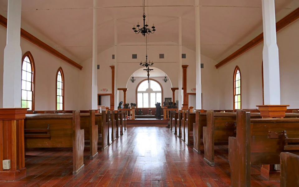 Interior of a church with wooden pews, chandeliers, arched windows, and a raised altar at the front.
