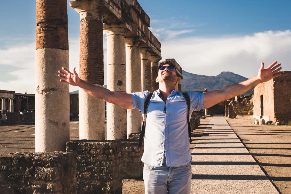 A person in sunglasses and casual clothing stands with arms open wide in front of ancient columns under a clear sky.