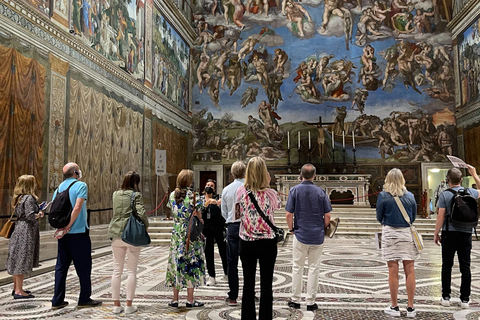 A group of people stand in the Sistine Chapel, admiring Michelangelo's ceiling frescoes and ornate wall decorations.