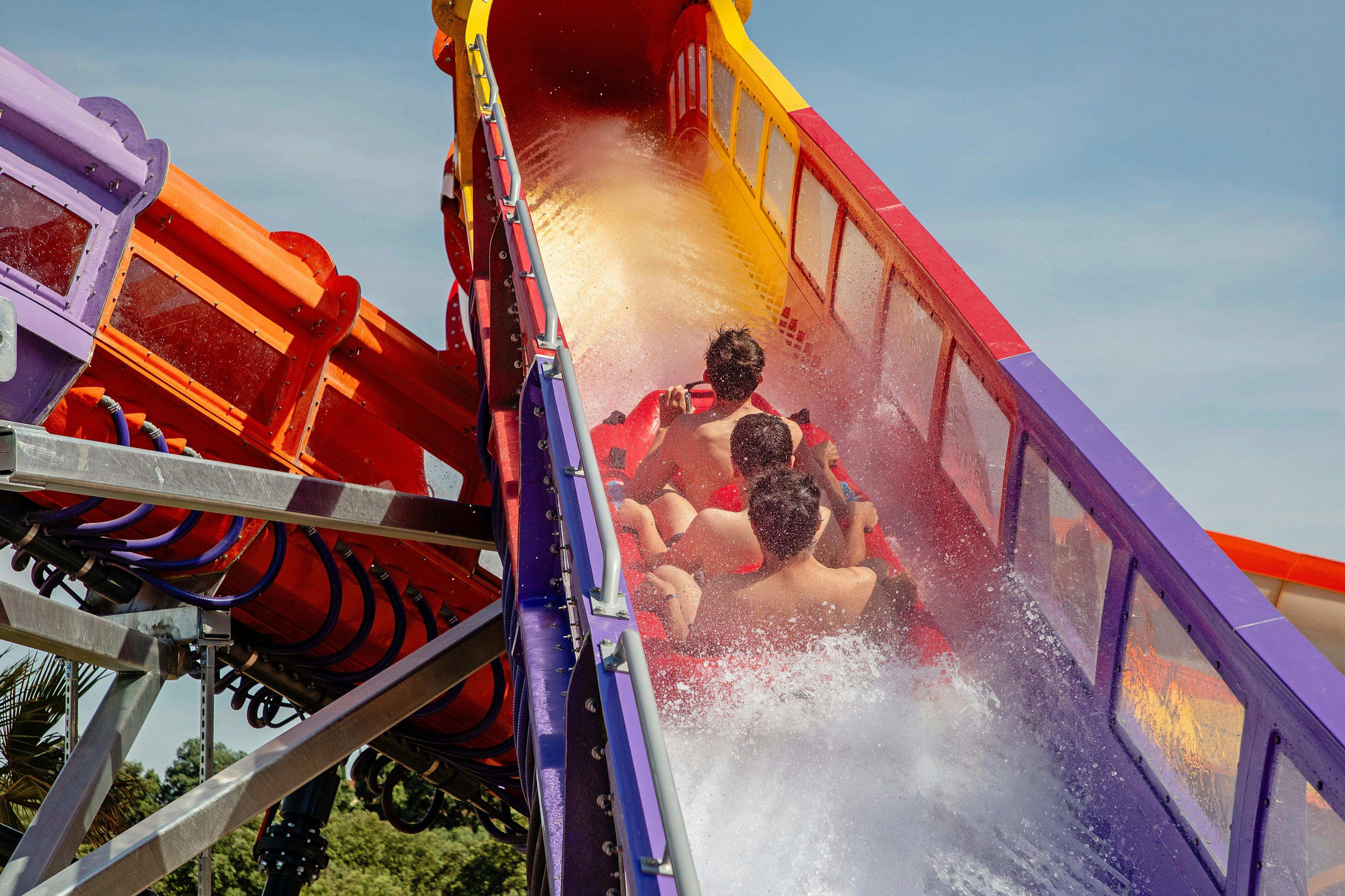 Un groupe de personnes sur un toboggan aquatique, éclaboussant l'eau, avec des sections du toboggan peintes en rouge, jaune et violet vifs.