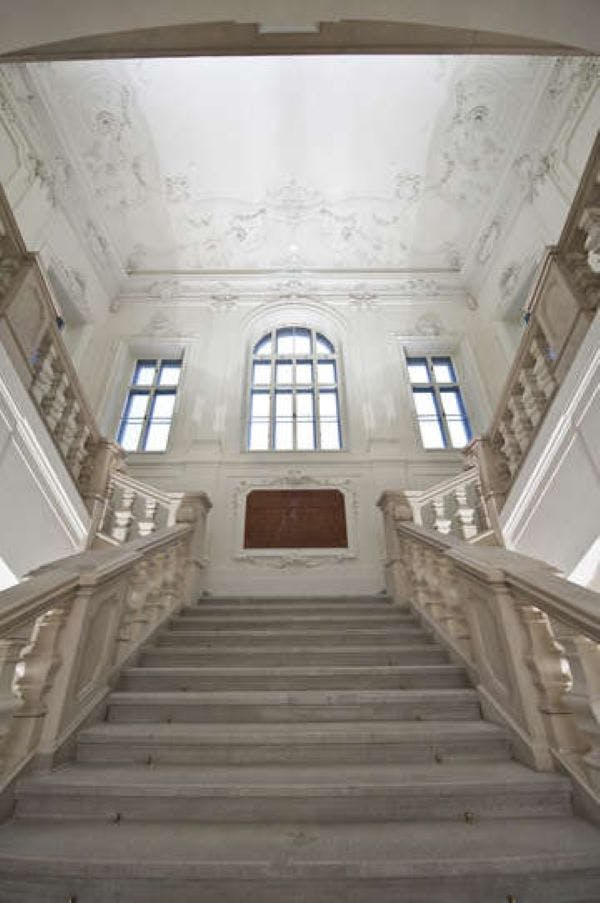A grand staircase with ornate railings leads to a landing with three large arched windows in an elegant, white interior.