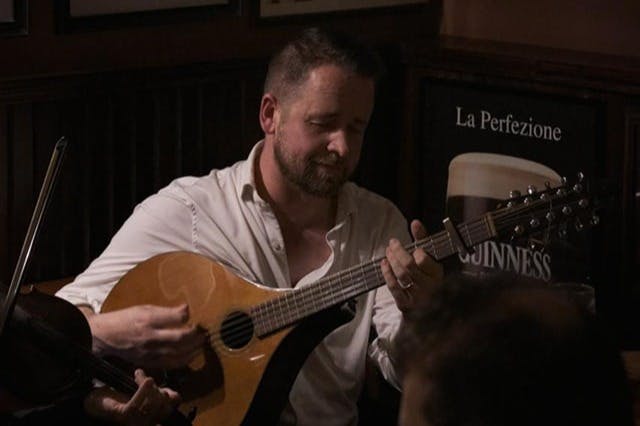 A person playing a guitar-like instrument in a dimly lit room with a relaxed expression. A Guinness poster is visible in the background.