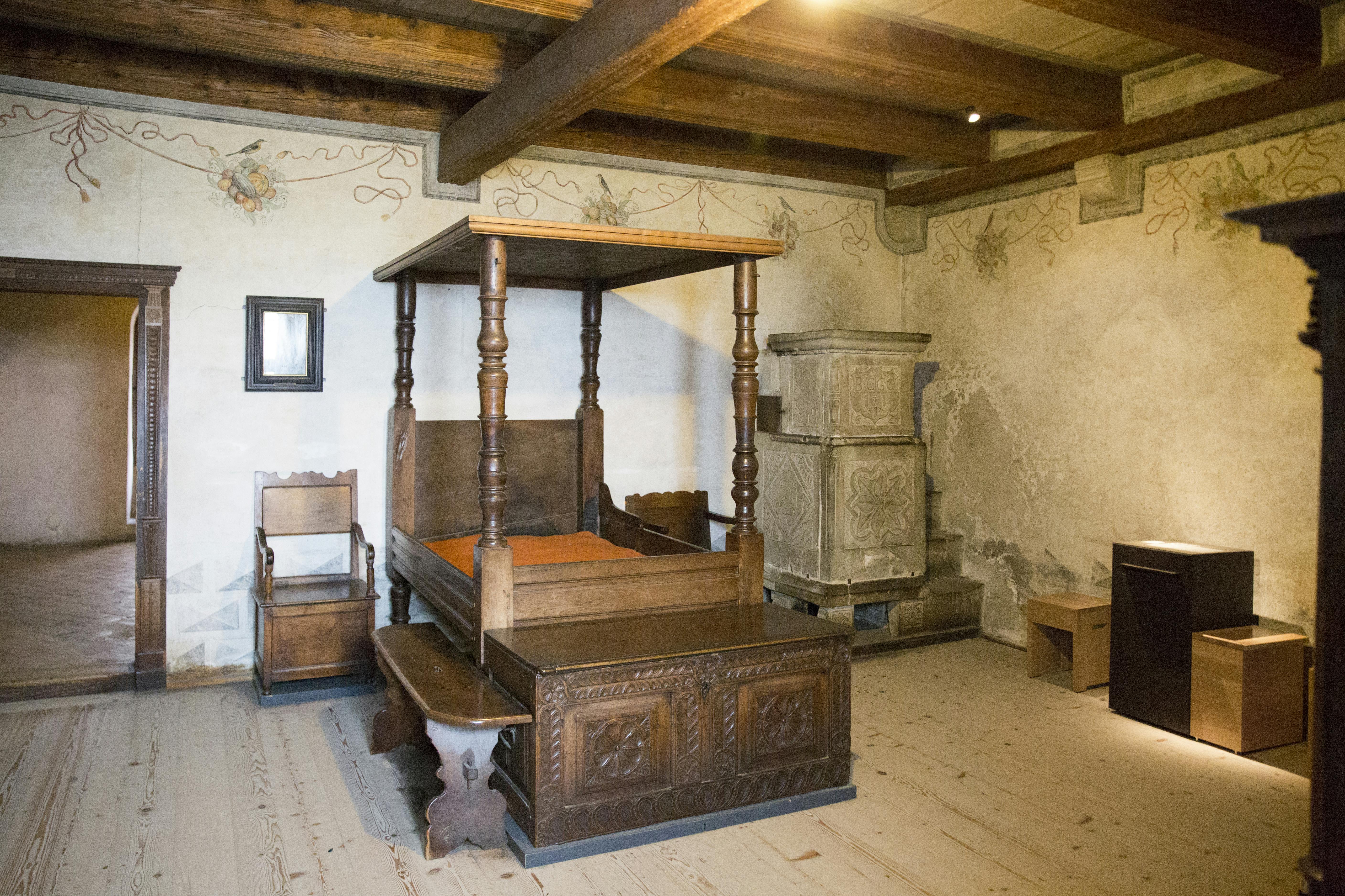 Room with a wooden four-poster bed, a carved chair, a bench, wooden beams on the ceiling, and an ornate ceramic stove.