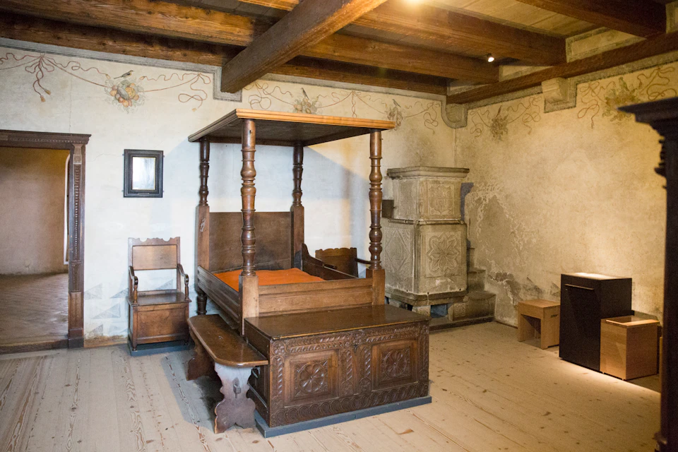 Room with a wooden four-poster bed, a carved chair, a bench, wooden beams on the ceiling, and an ornate ceramic stove.