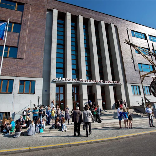 En grupp människor samlades utanför en byggnad med en "Národní Technické Muzeum" -skylt, en röd, vit och blå flagga och en trästruktur.