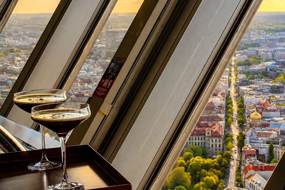 Two glasses on a table near a slanted window, overlooking a sunny cityscape with buildings, trees, and streets.