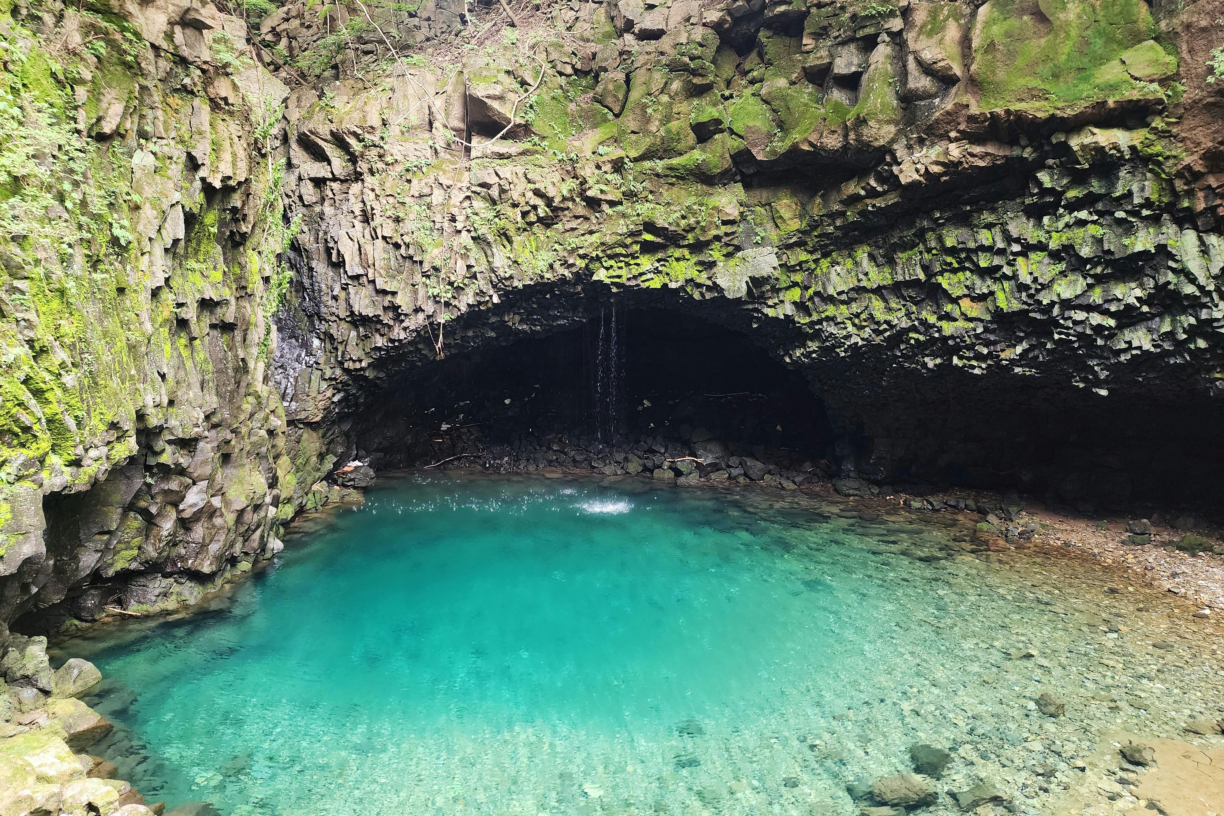 A turquoise pool of water beneath a rock overhang with moss-covered walls and a small waterfall in a natural setting.