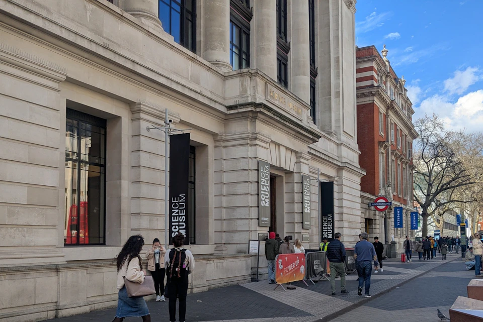 People walking in front of the Science Museum entrance in London, with banners and a red underground sign visible.