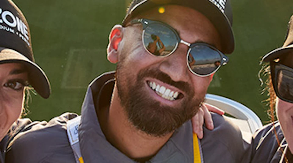 Two people wearing gear and "The OZONE" caps smile atop a stadium at sunset, with an empty field and city skyline in the background.
