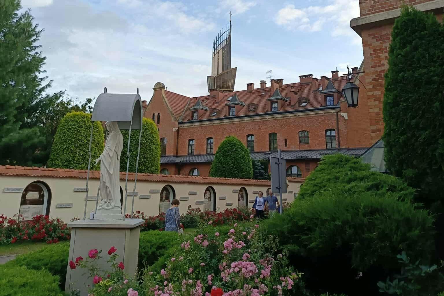 En trädgård med färgglada blommor, en staty och människor som går. En stor tegelbyggnad med ett högt torn är i bakgrunden.
