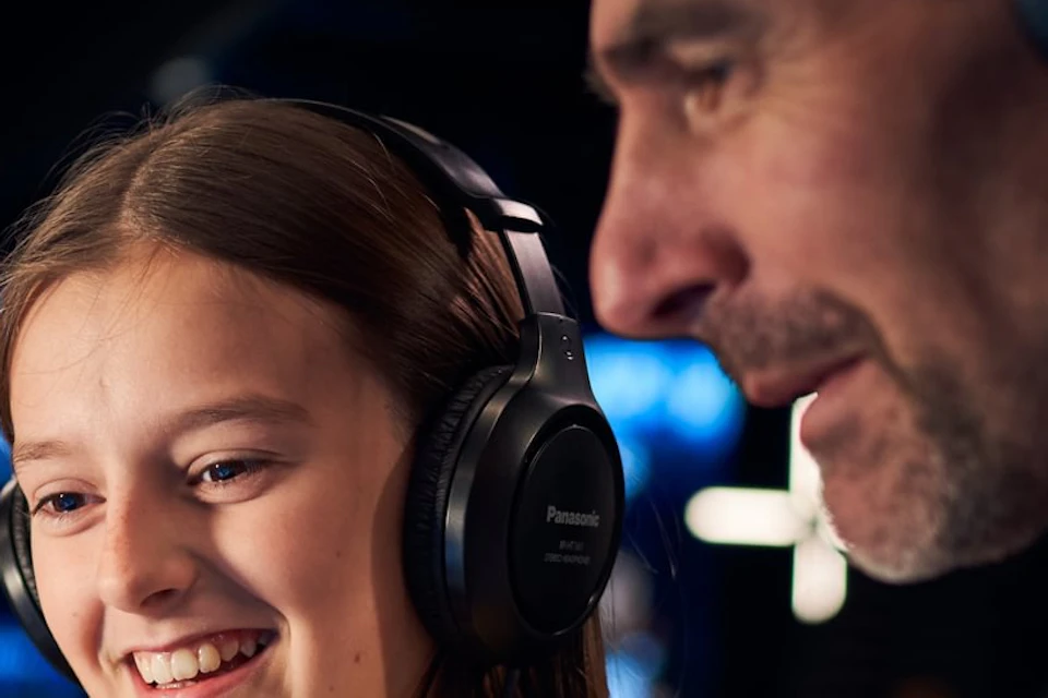 A man and a young girl wearing headphones interact with a touchscreen device in a dimly lit environment.