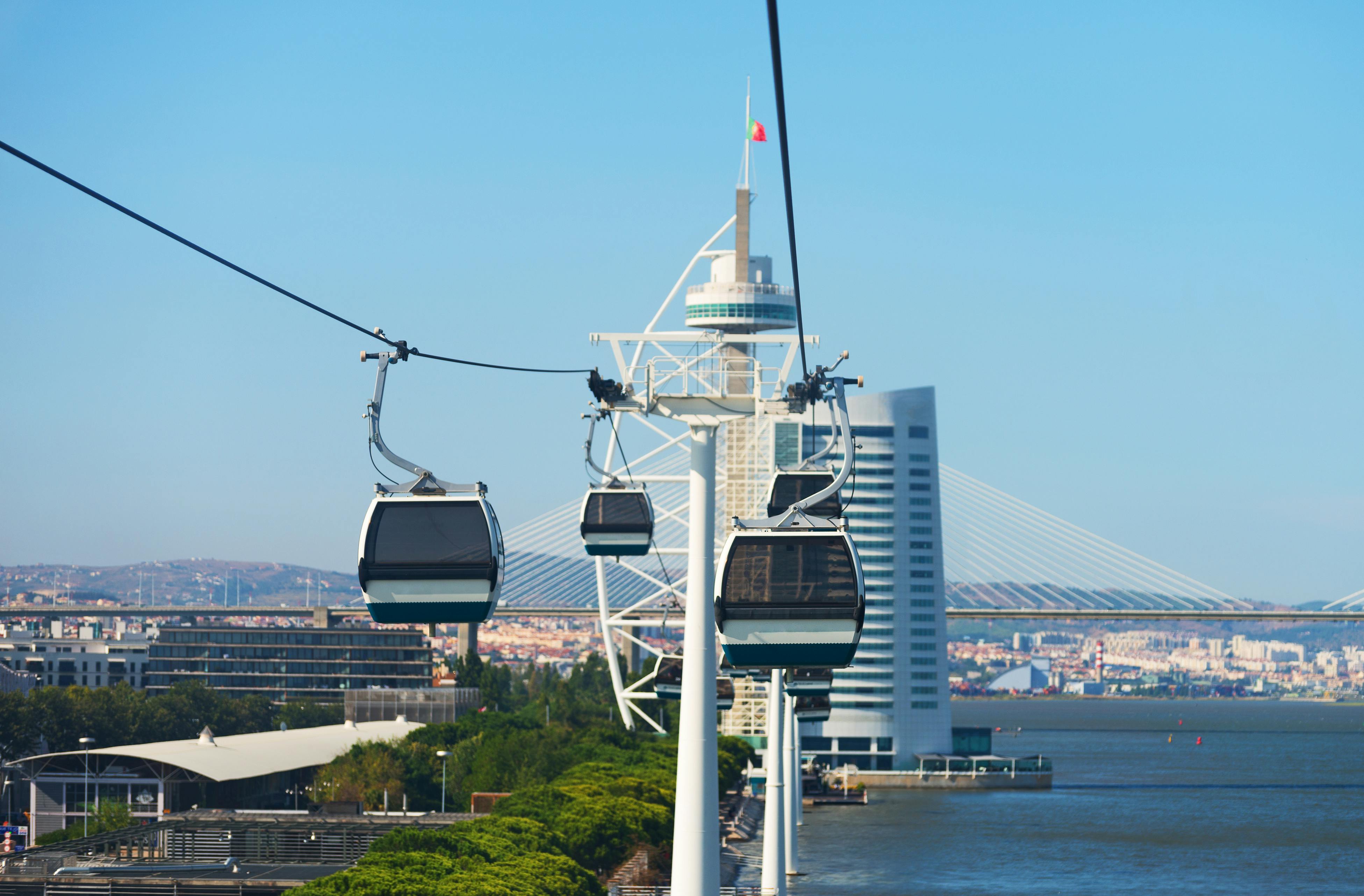Kolejki linowe nad rzeką z nowoczesną wieżą, mostem i pejzażem miejskim w tle pod czystym błękitnym niebem.