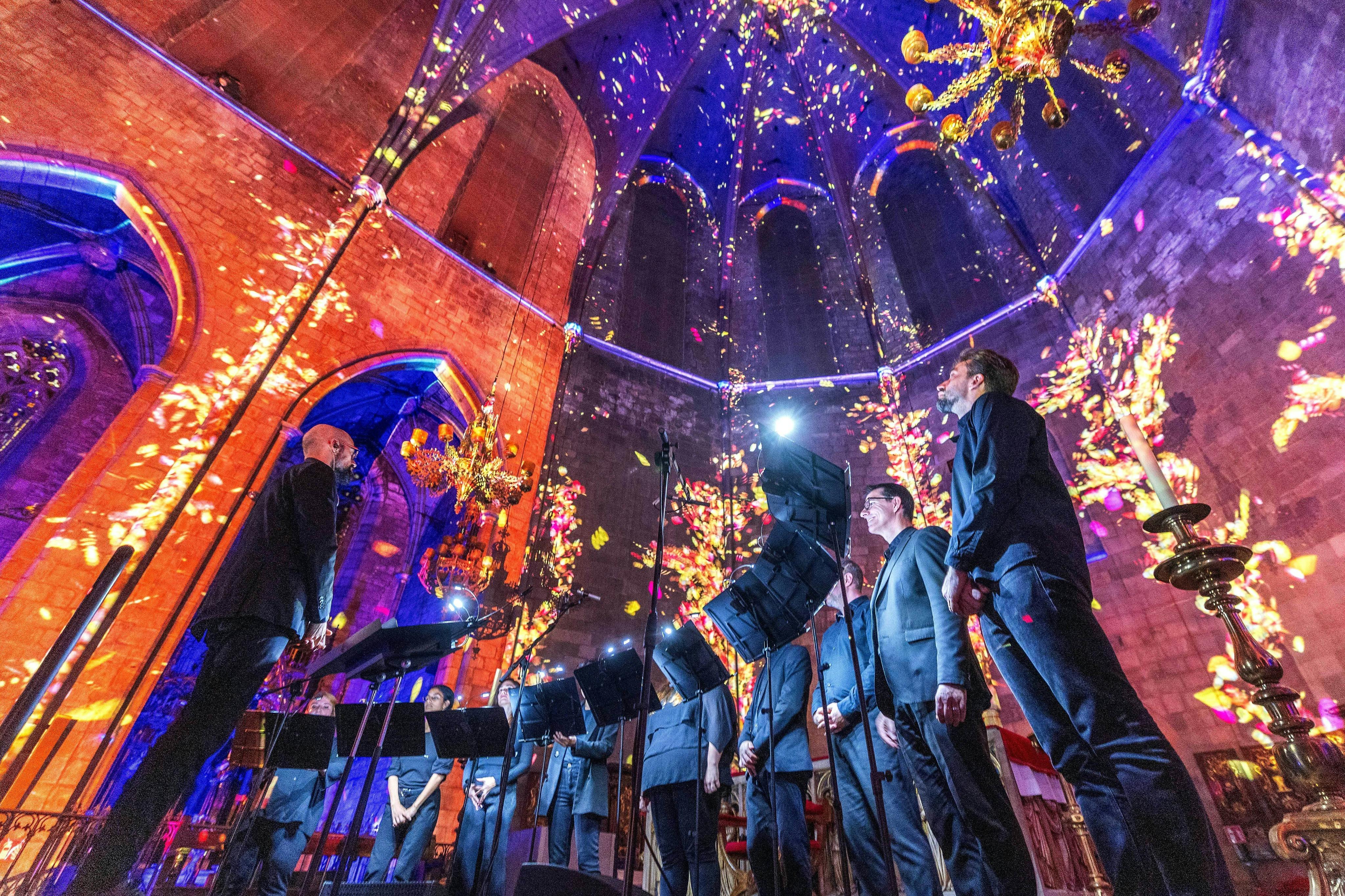Des choristes se produisent dans une cathédrale dont les murs et le plafond sont ornés de projections lumineuses colorées et florales.