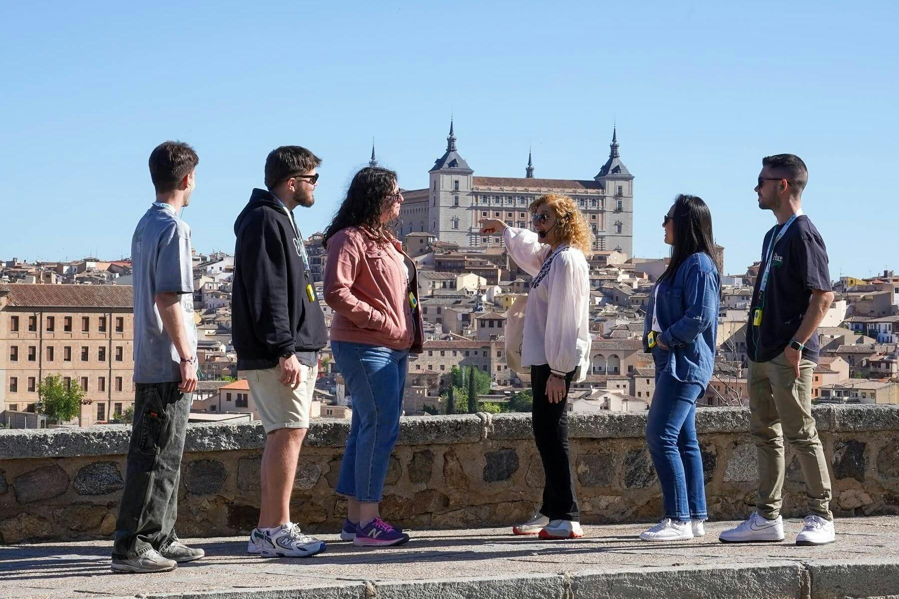Four people stand on a stone pathway overlooking a historic city with a prominent building and clear blue sky.