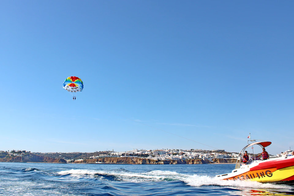 Two people parasailing over the water attached to a colorful parachute, with a boat and a coastal town in the background.