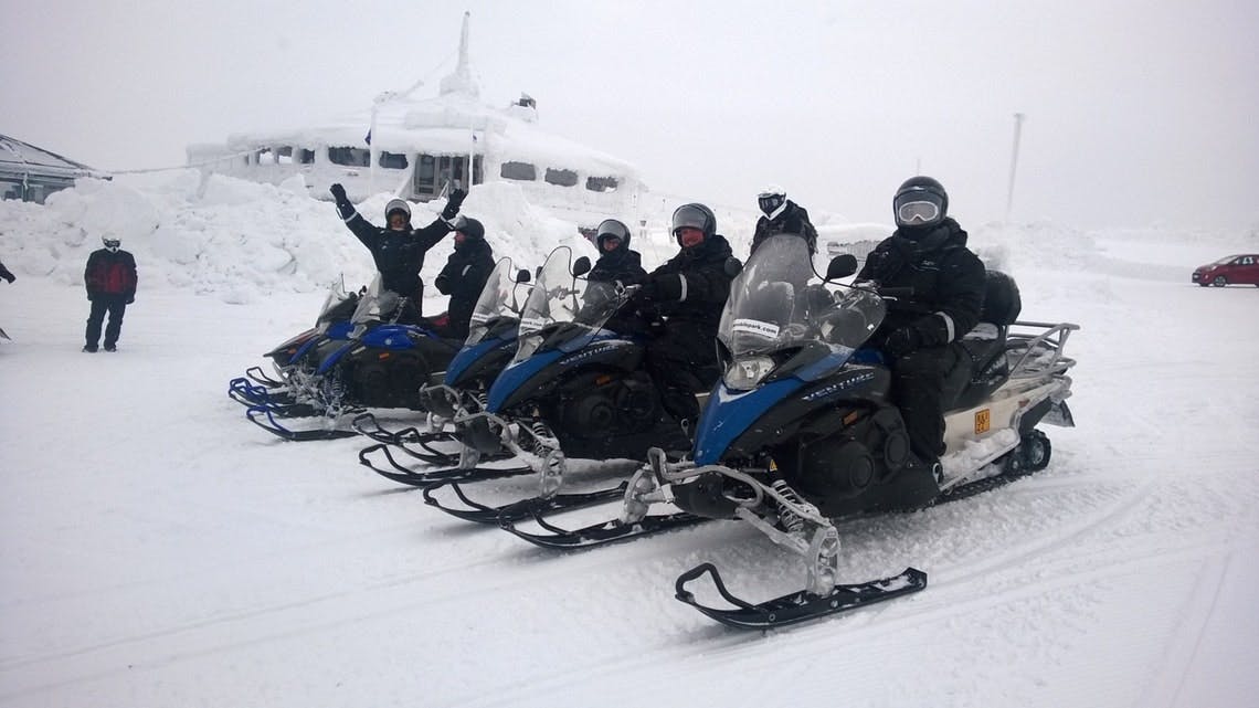 Πέντε άνθρωποι με χειμερινό εξοπλισμό κάθονται σε μπλε και λευκά snowmobiles σε ένα χιονισμένο τοπίο, με ένα χιονισμένο κτίριο στο παρασκήνιο.