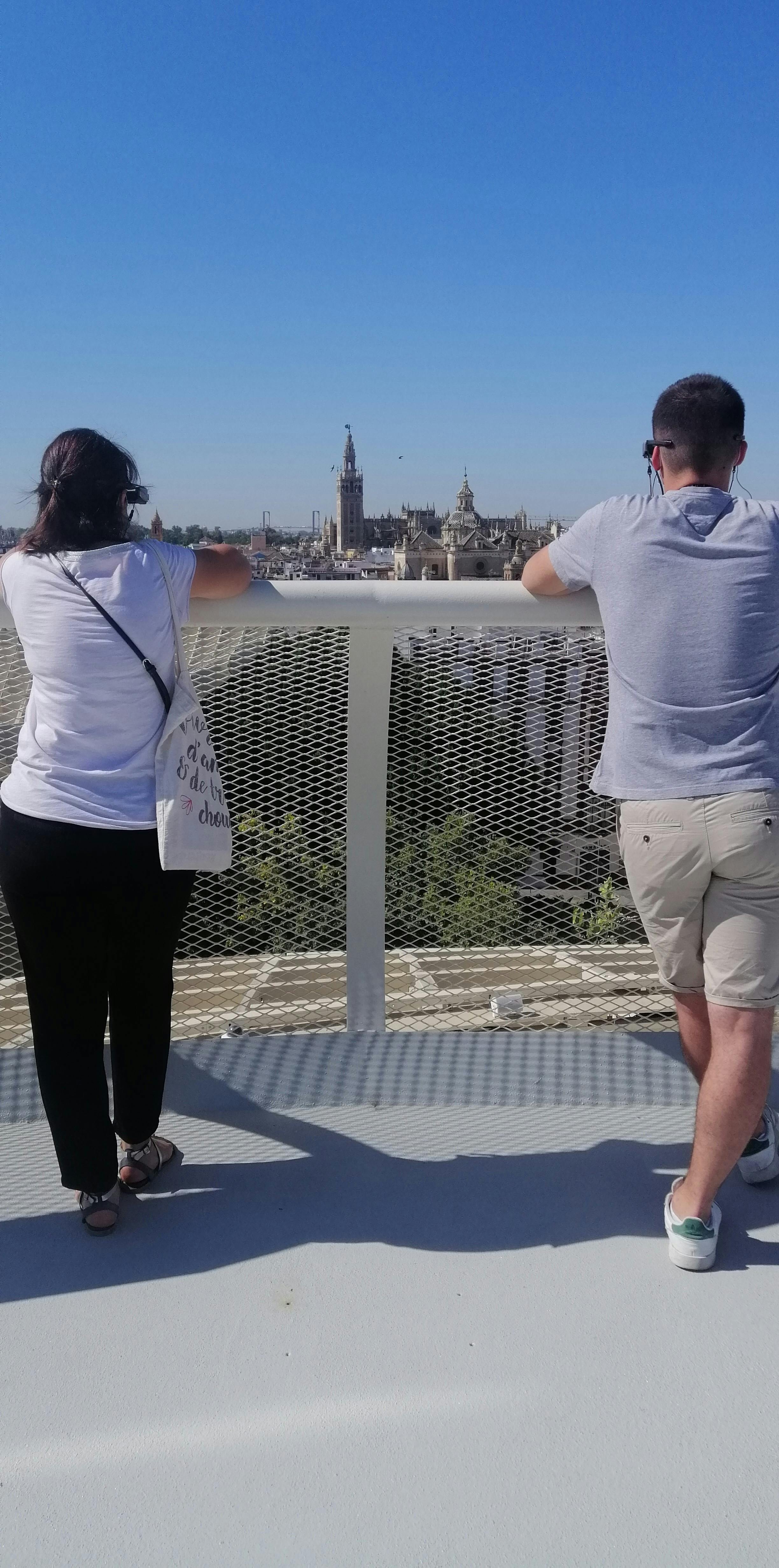 Δύο άτομα στέκονται σε ένα μπαλκόνι, με θέα ένα αστικό τοπίο με έναν προεξέχοντα πύργο στο παρασκήνιο. Καθαρός γαλάζιος ουρανός από πάνω.