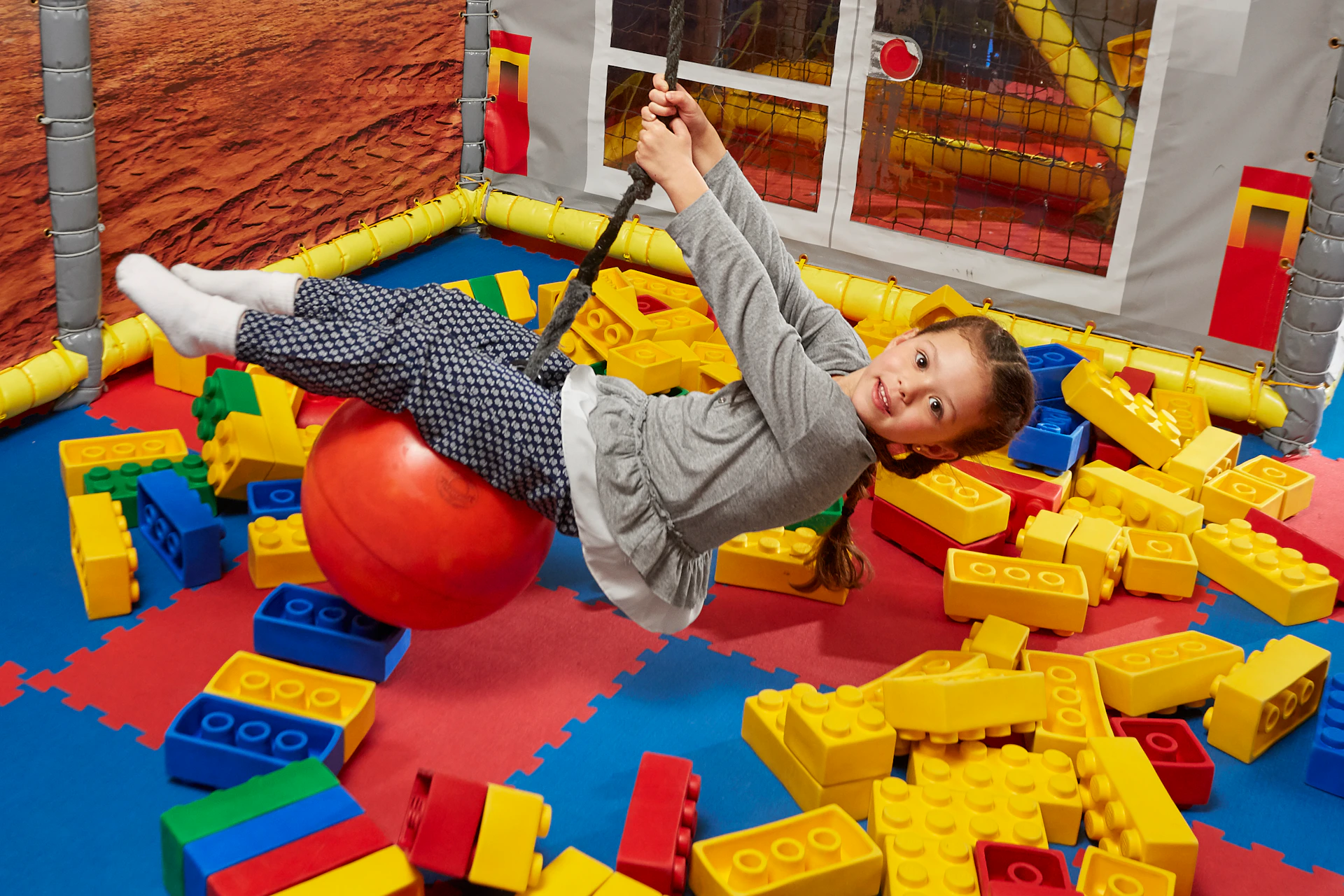 A young girl swings on a red ball in an indoor play area filled with large yellow and red building blocks.