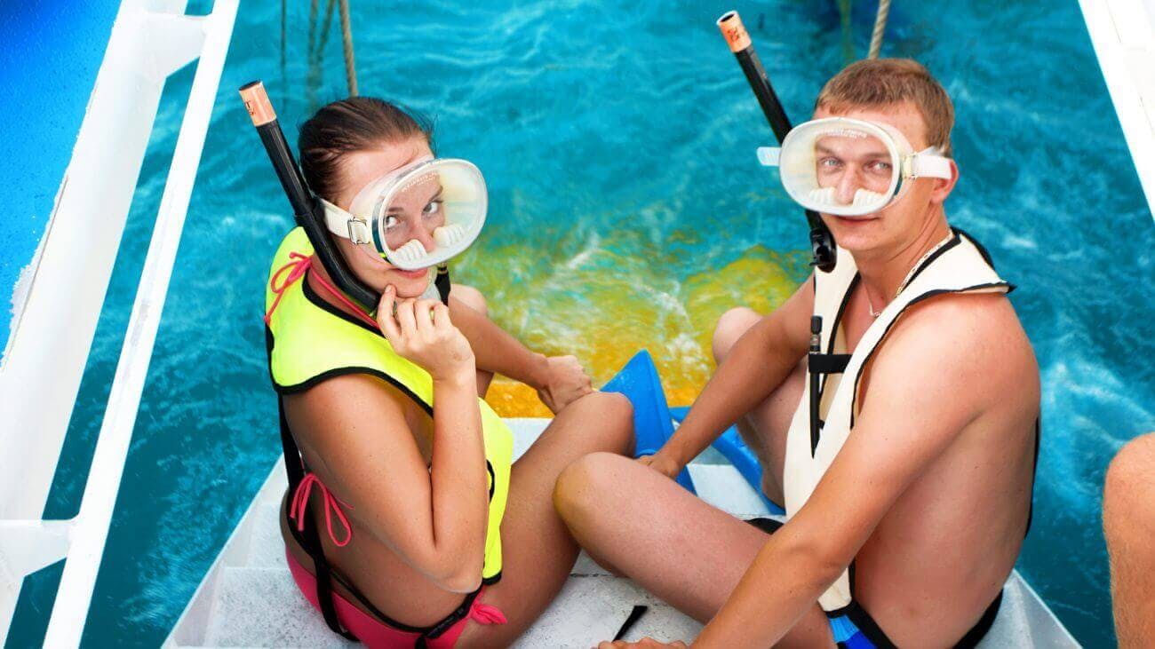 Muž v masce šnorchlu a záchranné vestě sedí na lodi u vody a je připraven šnorchlovat. Je vidět modrá voda a část jiné osoby.