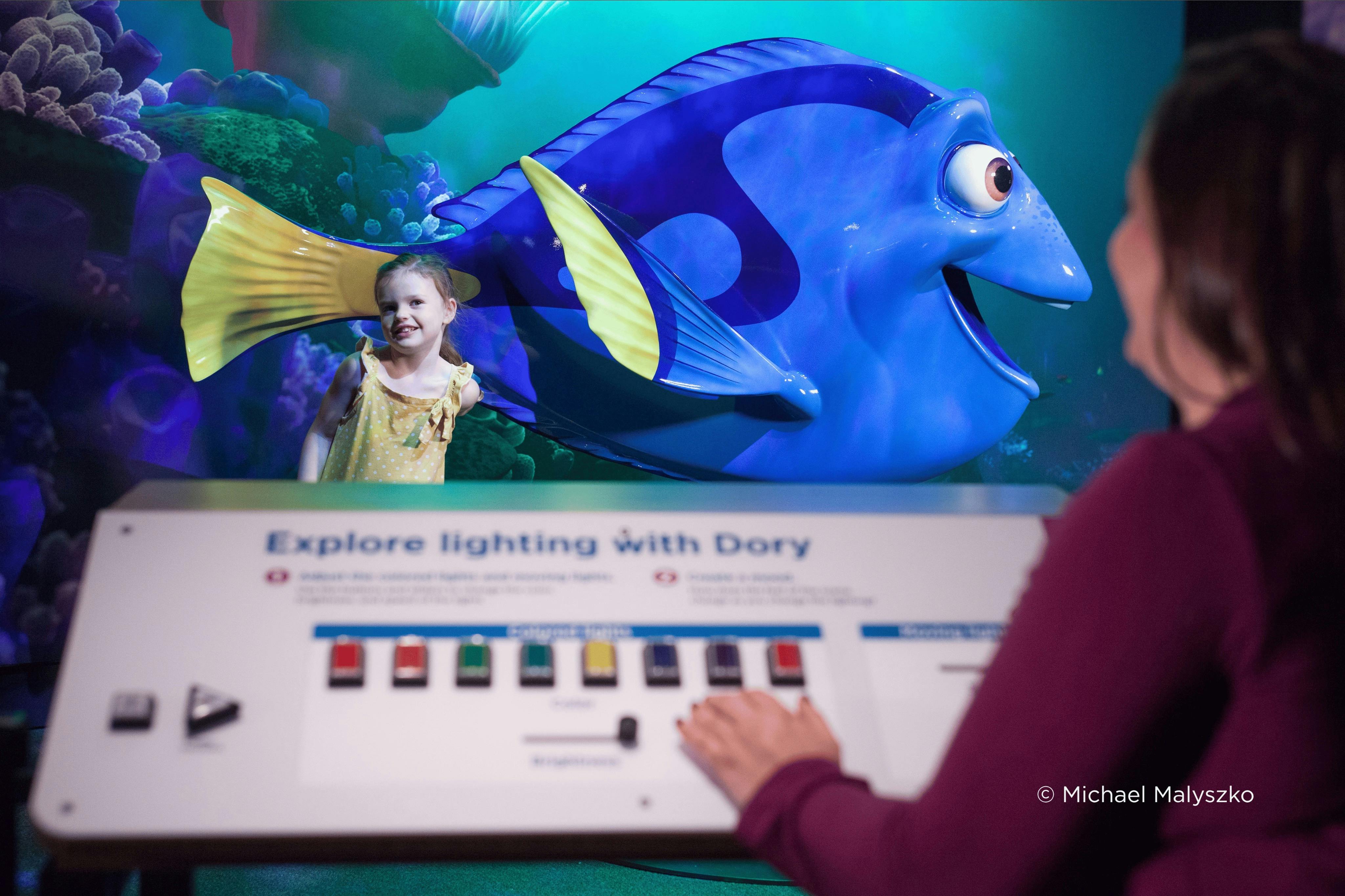 Un enfant interagit avec une installation sur le thème de l'univers aquatique, comprenant un grand aquarium et un panneau de commande intitulé « Découvre l'éclairage avec Dory ».