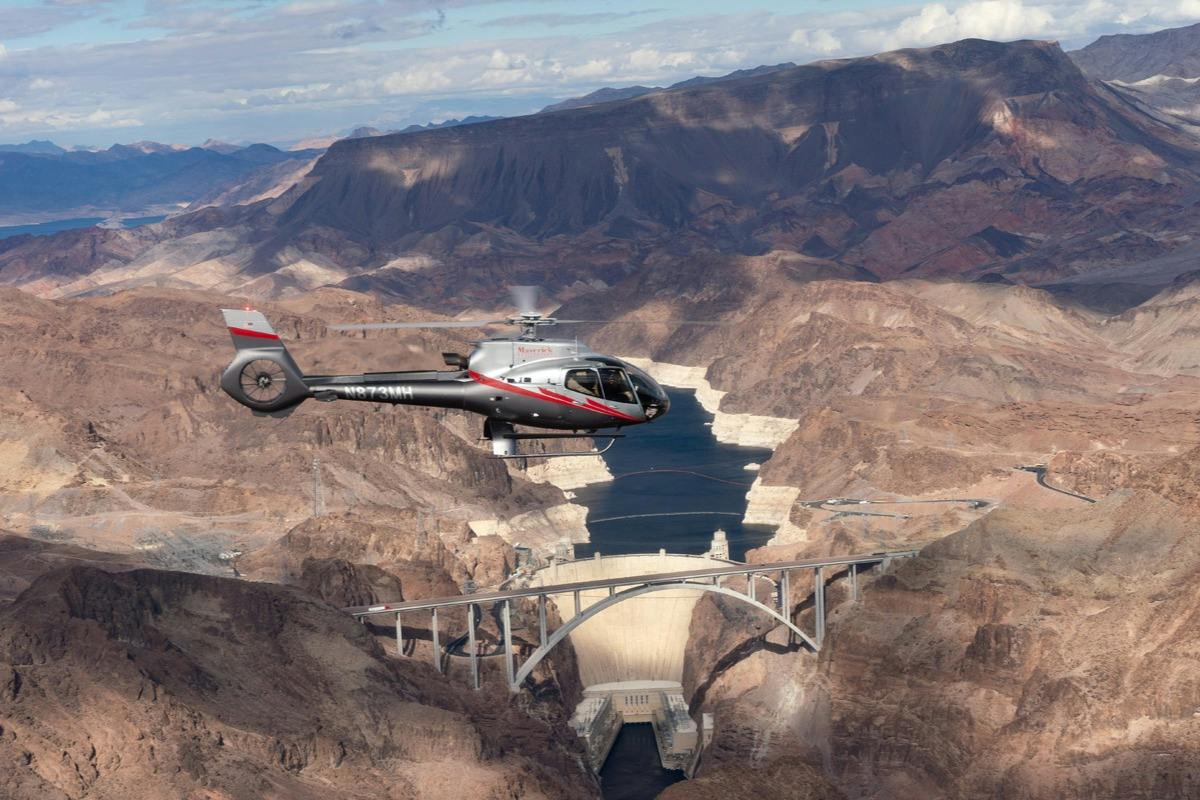 A helicopter flying over a dam and a bridge in a desert canyon under a partly cloudy sky.