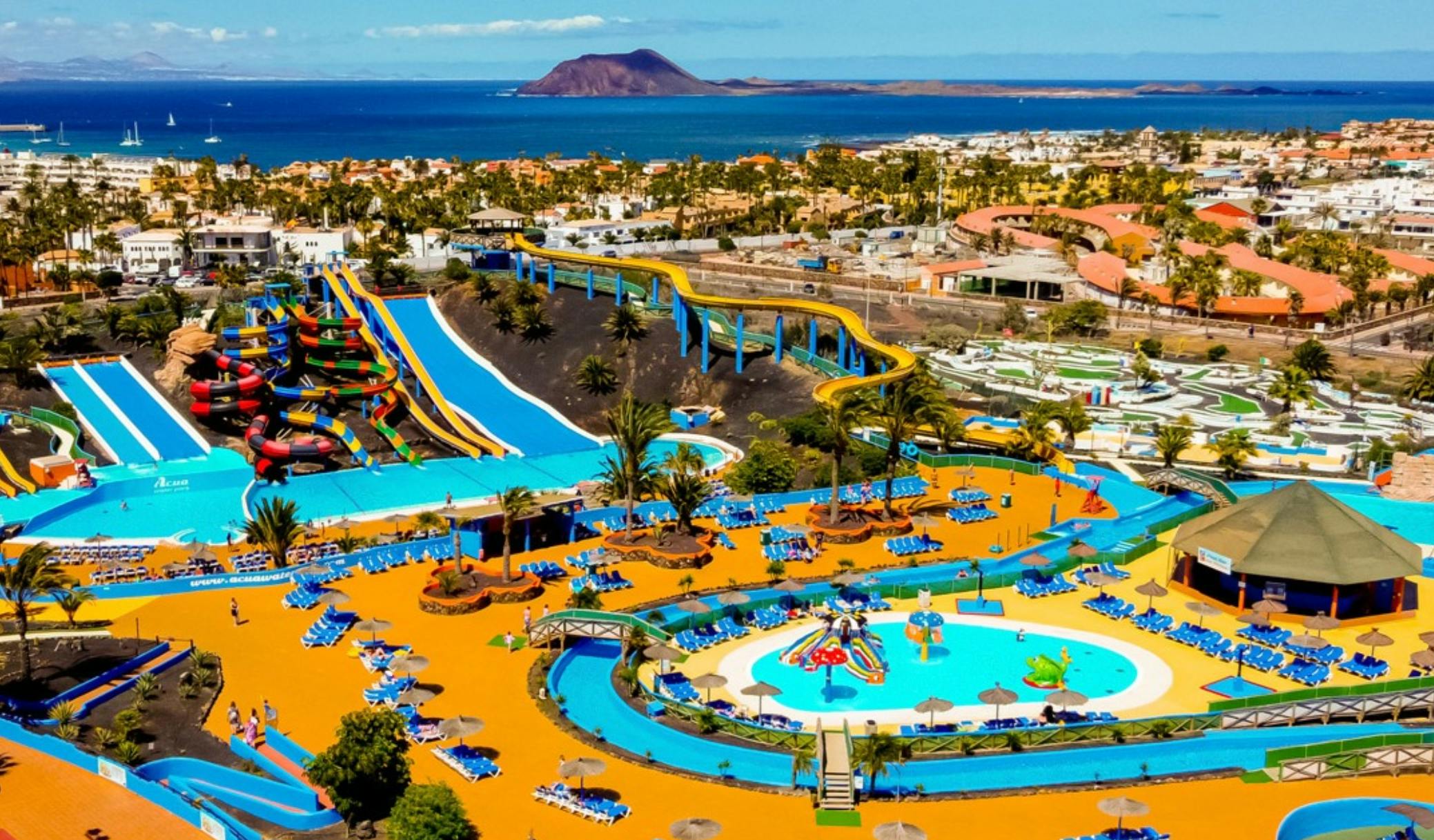 Een kleurrijk waterpark met zwembaden, glijbanen, lounges en groen, gelegen bij een kustplaats met uitzicht op de oceaan.