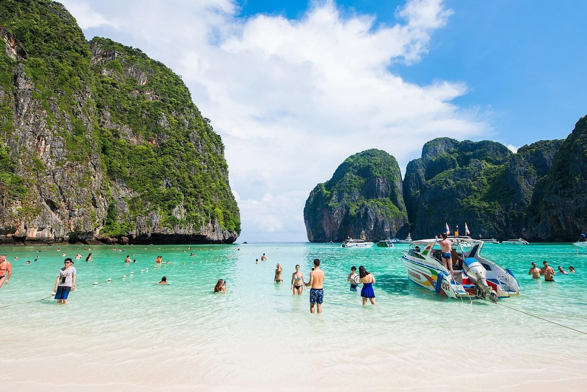A tropical beach with clear turquoise water, people swimming, and boats near limestone cliffs under a blue sky.