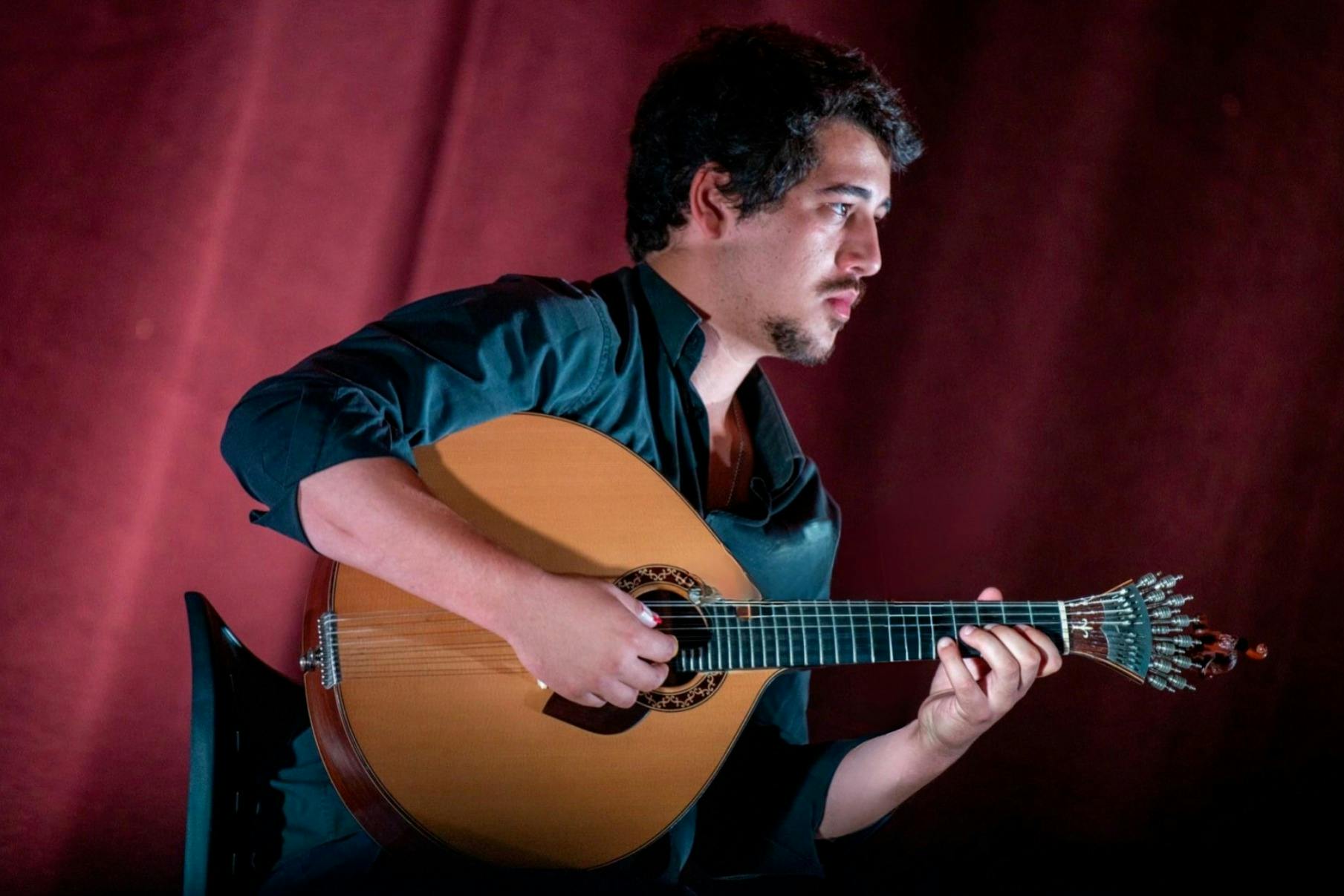 Ένα άτομο με σκούρα μαλλιά και γένια, που φορά μαύρο πουκάμισο, παίζει ένα έγχορδο όργανο σε σκούρο κόκκινο φόντο.