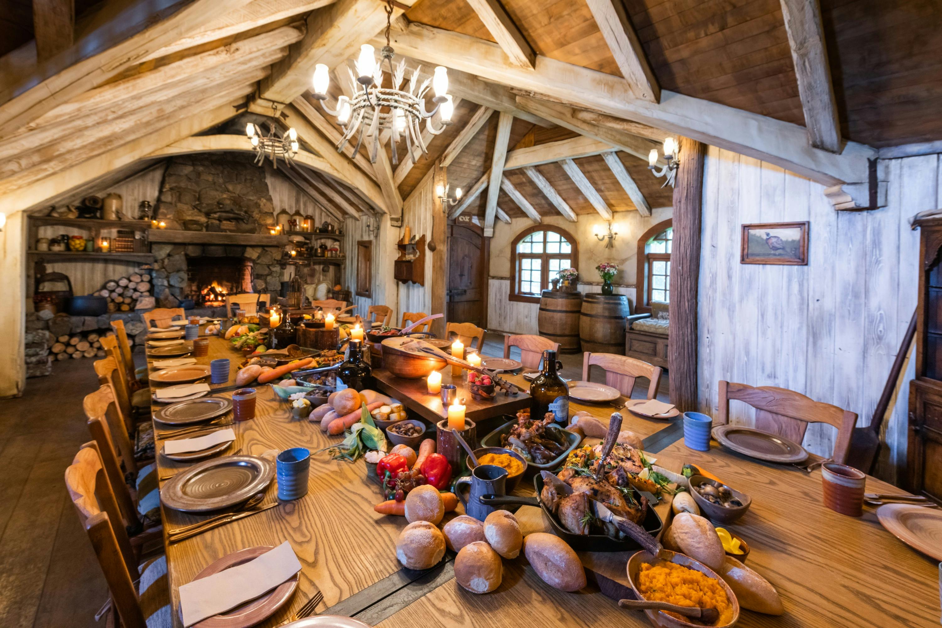 A rustic dining room with a long wooden table set for a feast, surrounded by wooden beams and warm lighting.