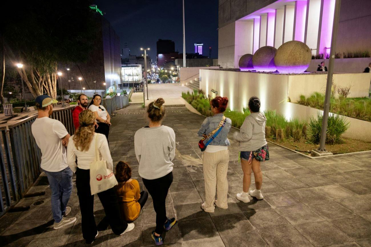 A group of people standing and sitting on a paved sidewalk at night, with lit buildings and trees in the background.