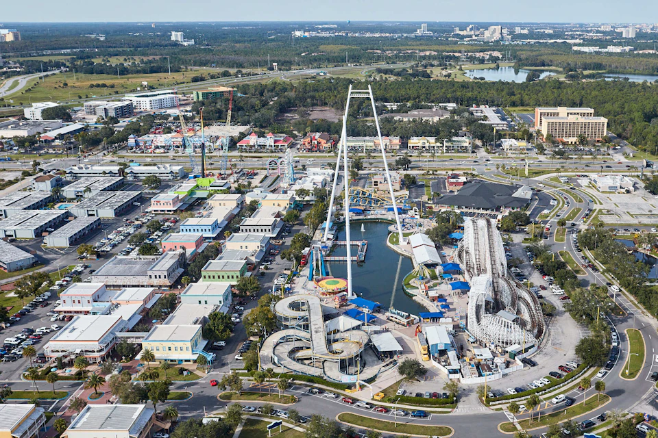 Aerial view of an amusement park with various rides, buildings, and green spaces surrounded by roads and distant cityscape.