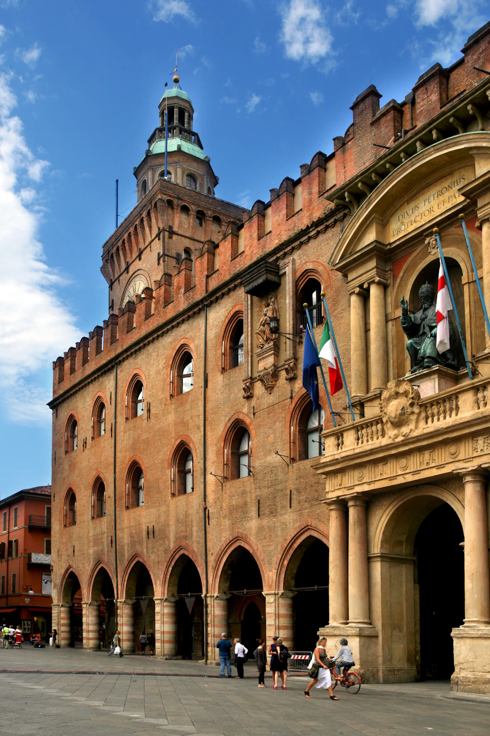 Historic brick building with arched windows, flags, and statues under a blue sky.