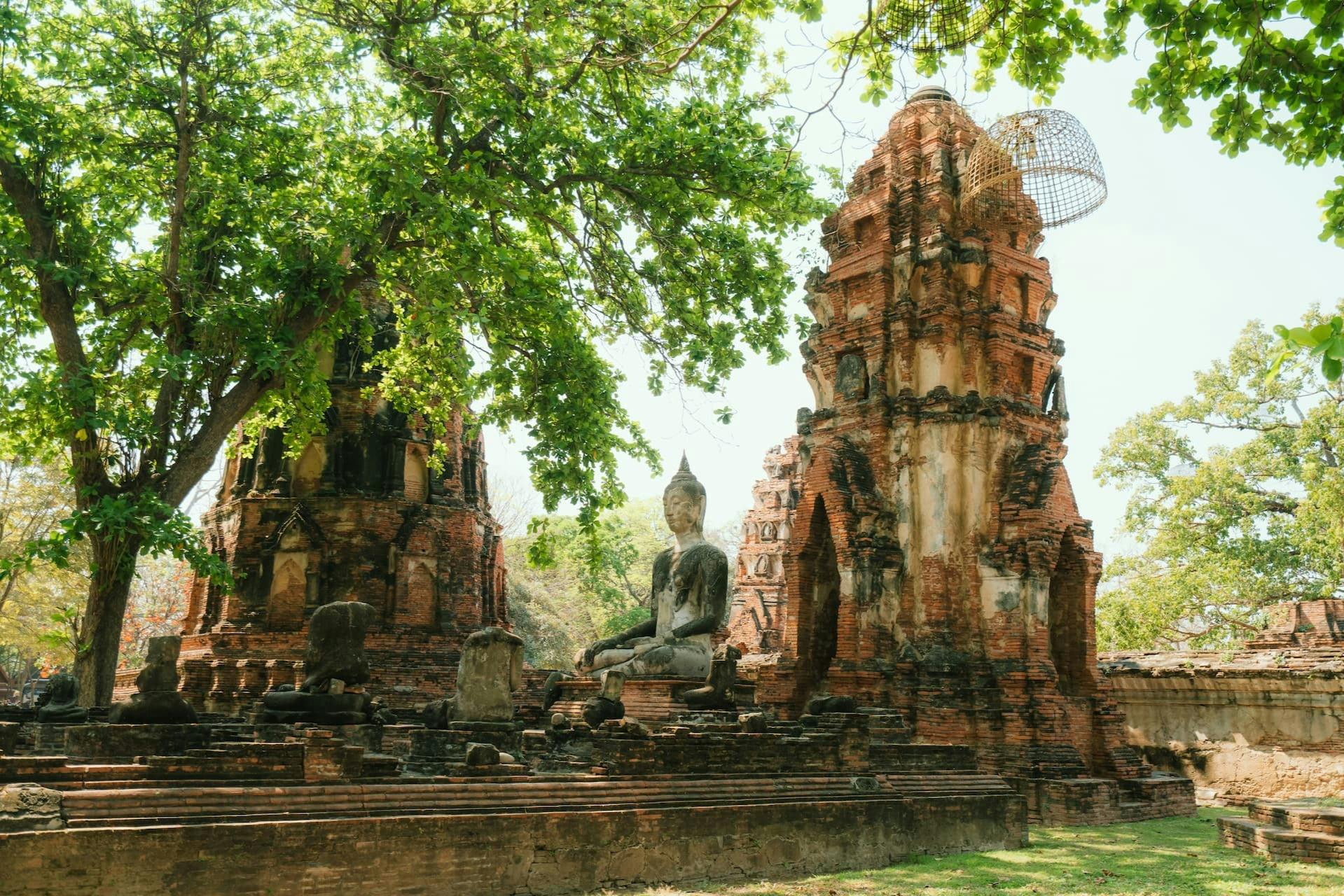 Gamle tempelruiner med en siddende Buddha-statue omgivet af træer under stærkt sollys.