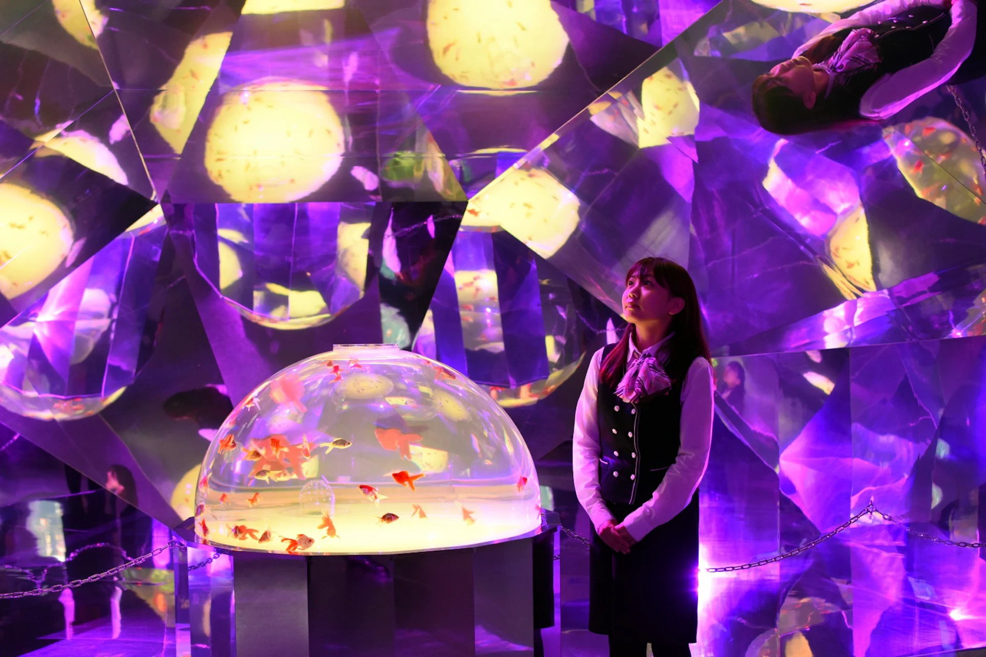 A woman stands beside a lit dome containing goldfish, surrounded by colorful reflective surfaces in an artistic display.