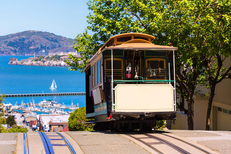 A cable car on a hill with blue tracks, overlooking a marina and water with a sailboat, framed by trees and distant hills.