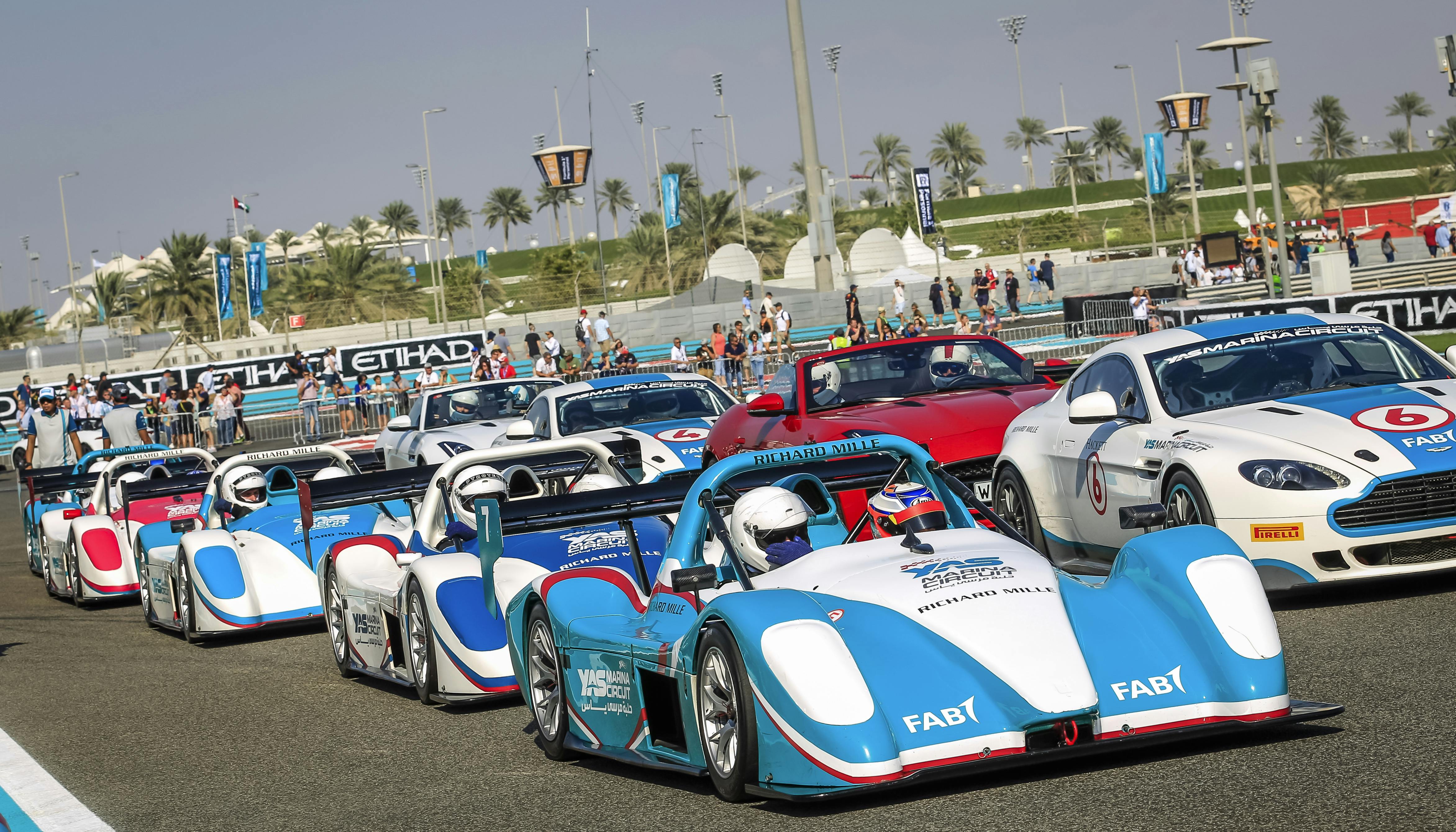 Race cars with drivers in helmets lined up at a starting line with spectators and palm trees in the background.