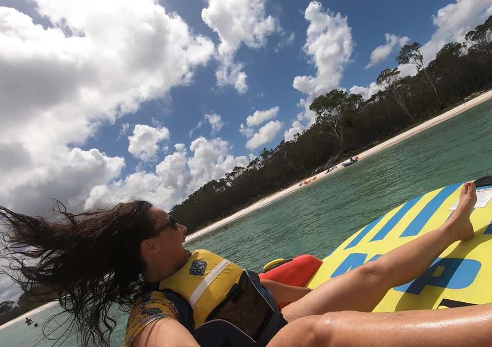 Person with a yellow life vest on a colorful inflatable raft in clear water, with a sandy beach and trees in the background.
